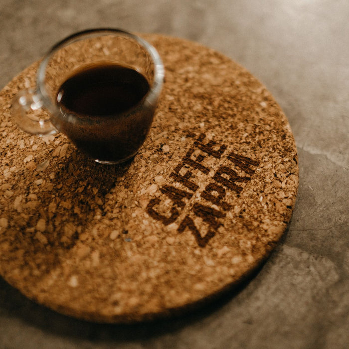 A cork coaster holding a glass of black coffee on a concrete surface.