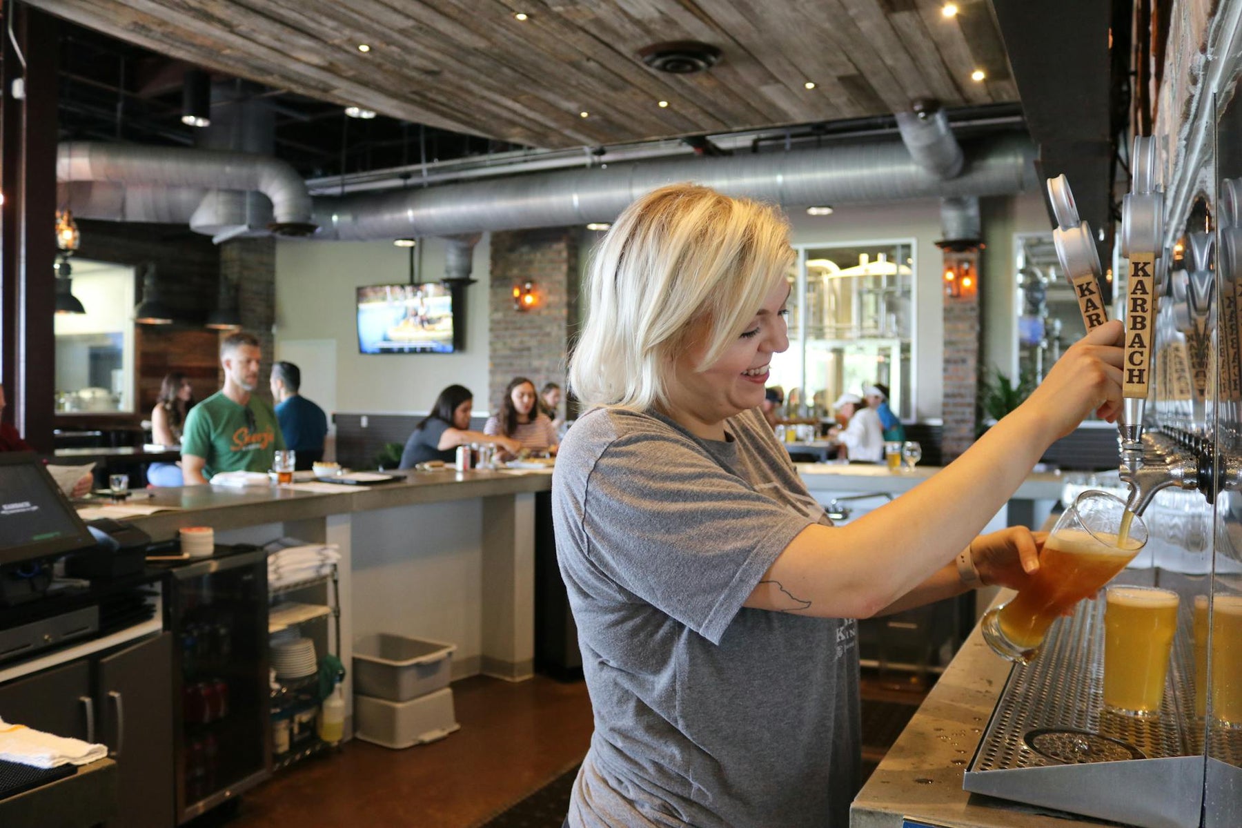 A smiling bartender pours a beer for customers in a lively, industrial-style bar setting.