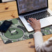 Desk Mat - A desk mat featuring a leaf pattern in green and white, with a logo design, beside a laptop and smartphone.