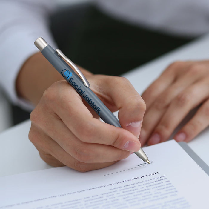 Lancer Fashion Pen - A metal pen in grey is being held, with a logo visible. A hand is writing on a sheet of paper.