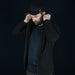 Swiss Peak Urban Hoodie - A man in a black Swiss Peak Urban Hoodie adjusts the hood, wearing a black cap. The background is dark.