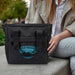 Oceana Recycled Cooler Bag - A black cooler bag with a logo sits beside a person wearing a light coat and jeans.
