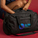 Oceana Recycled Duffle Bag - A black duffle bag with handles and a logo, held by a person, set against a red background.