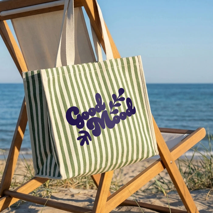 Keepsake Beach Tote Bag - A green and white striped tote bag with a branding logo sits on a wooden beach chair, with a scenic ocean view in the background.