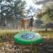 Aero Flyer - Small - A green frisbee rests on the ground, surrounded by trees and people playing in the background. It features a logo.