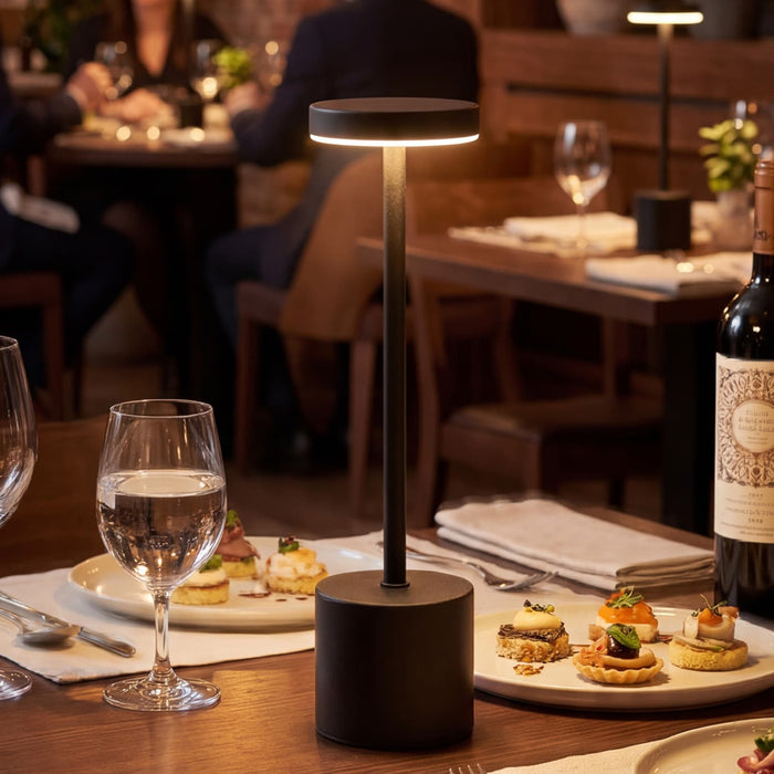Nouveau Rechargeable Table Lamp - A black rechargeable table lamp stands on a dining table, illuminating an elegant setting with food and drinks.