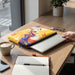 Full Colour Laptop Sleeve - Medium - A medium laptop sleeve featuring a vibrant yellow design with abstract patterns, resting on a wooden table beside a coffee cup.