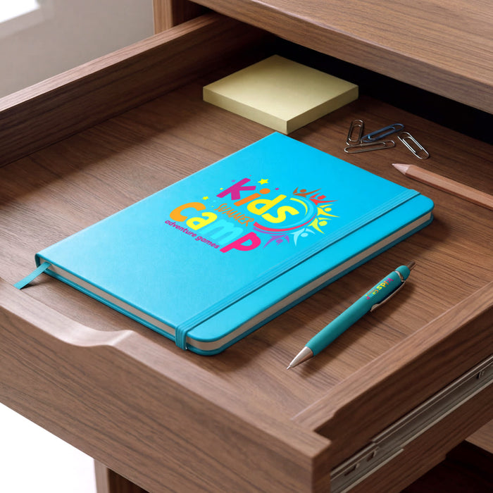 Omega Notebook and Pen Gift Set - A blue notebook with a colourful design and a turquoise pen are placed on a wooden desk drawer.