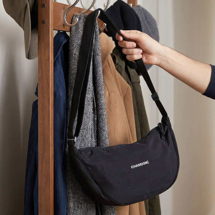 Selene Moon Bag - A black shoulder bag is being held on a coat rack, surrounded by jackets and hats. The bag has a logo.
