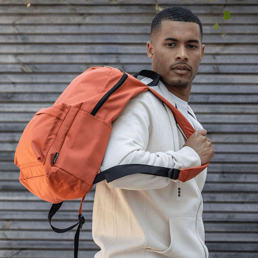 Byron 14" GRS Recycled City Laptop Backpack 16L - A man in a white hoodie carries an orange laptop backpack, featuring multiple compartments and a logo.