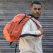 Byron 14" GRS Recycled City Laptop Backpack 16L - A man in a white hoodie carries an orange laptop backpack, featuring multiple compartments and a logo.
