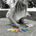 STAEDTLER Assorted Sidewalk Chalk Set - A child uses colourful chalk sticks, including white, orange, pink, green, and purple, from an open box on the ground.