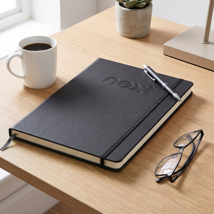 A4 Daily Diary - 2027 - A black A4 diary laid on a wooden desk, accompanied by a pen, glasses, a coffee cup, and a small plant.