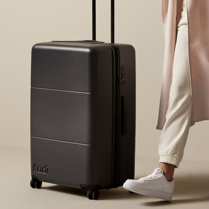 Kadi Check-In Suitcase - Large - A large black suitcase on wheels with a retractable handle and a logo.