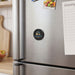 Multi-Functional Magnetic Badge - A round black magnetic badge with a logo attached to a stainless steel fridge, alongside a notepad and groceries.