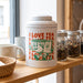 Prism Print - 100 x 100mm Sticker - A white tea canister with green illustrations of two cheerful mugs, displayed on a wooden shelf alongside jars and cups.