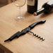 Premium Wine Waiters Knife - A black wine waiters knife positioned on a wooden table, alongside a wine bottle and a glass.