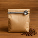 Coffee Measuring Spoon With Clip - A brown paper bag of coffee with a stainless steel measuring spoon resting on top, surrounded by coffee beans.