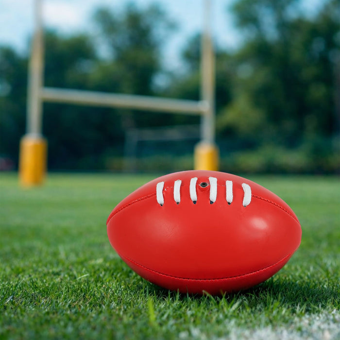 Mini Promotional Aussie Rules Ball - A red mini promotional Aussie Rules ball resting on green grass, with goalposts blurred in the background.