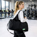 Duffle Bag With Yoga Mat Strap - A black duffle bag with a strap, carried by a woman in a gym setting. It features a logo on the side.