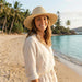 Airlie Paper Straw Hat - A straw hat in natural tones, worn by a woman on a beach, surrounded by palm trees and water.
