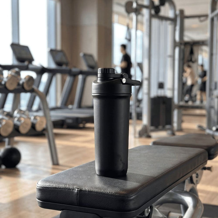 Forge Double Wall Shaker 750ml - A drink bottle with a double-wall design in black, placed on a gym bench with exercise equipment in the background.