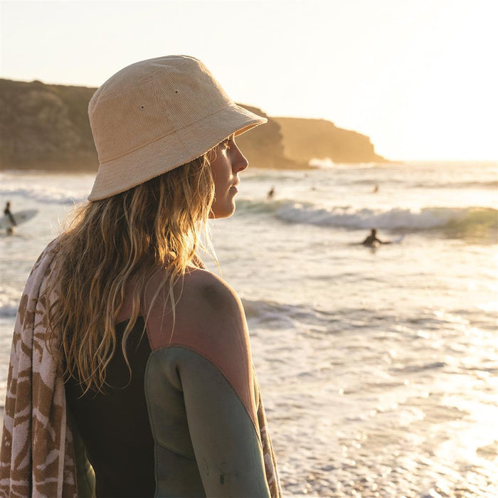 Leroy Cord Bucket Hat - A woman stands by the ocean in a beige bucket hat, with surfers visible in the background during sunset.