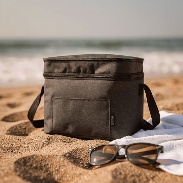 Tundra GRS RPET Double Compartment Cooler Bag 13L - A grey double compartment cooler bag sits on sandy beach alongside sunglasses and a towel, with ocean waves in the background.