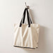 Sierra Foldable GRS Recycled  Cotton Tote Bag - A beige foldable tote bag with black straps hanging on a hook. It features a logo.
