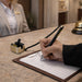 Counter Fixed Signature Pen E - A sleek black and gold pen is being used to sign a document on a reception desk with a gold holder.
