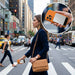 Apex Semi-Solid-State 22.5W MagSafe Wireless Power Bank - 5000 mAh (Stock) - A woman in a navy suit walks across a city street, holding an orange MagSafe power bank and smartphone.
