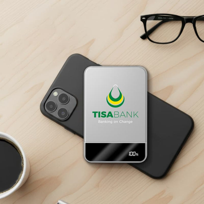 Apex Semi-Solid-State 22.5W MagSafe Wireless Power Bank - 5000 mAh (Stock) - A silver MagSafe wireless power bank resting on a black phone, beside a coffee cup and glasses.