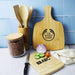 Tiga Bamboo Cutting Board - A bamboo cutting board in natural colour, alongside fresh vegetables, utensils, and a jar of ingredients. Features a logo.