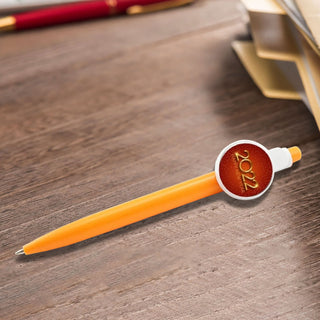 Button Badge Pen - An orange plastic pen with a circular badge on the side, resting on a wooden surface beside stationery.