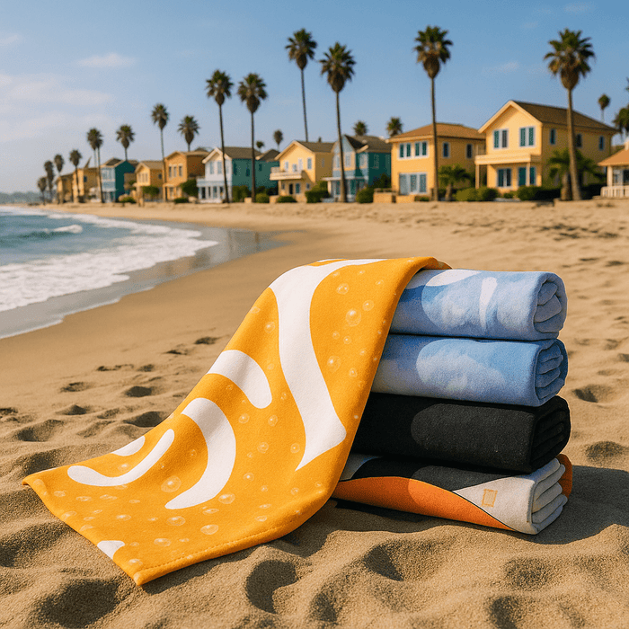 Bath Towel - A stack of five bath towels in various colours, including yellow, blue, and black, on a sandy beach with palm trees and houses in the background.