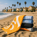 Bath Towel - A stack of five bath towels in various colours, including yellow, blue, and black, on a sandy beach with palm trees and houses in the background.