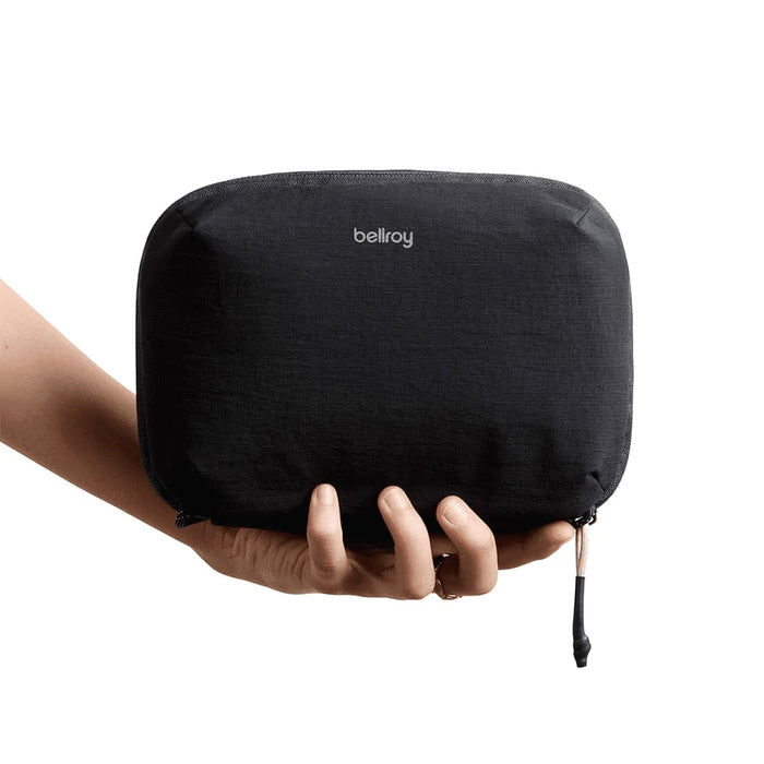 Bellroy Lite Tech Kit - A hand holding a black tech kit with a smooth texture and a logo. The zippered pouch is compact and sleek.