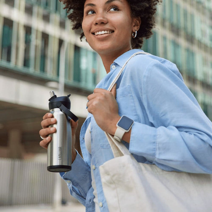 CamelBak - Thrive Flip Straw VSS 0.6L - A drink bottle in stainless steel, held by a person wearing a blue shirt and carrying a tote bag. The bottle features a flip straw and has a logo.
