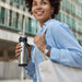 CamelBak - Thrive Flip Straw VSS 0.6L - A drink bottle in stainless steel, held by a person wearing a blue shirt and carrying a tote bag. The bottle features a flip straw and has a logo.