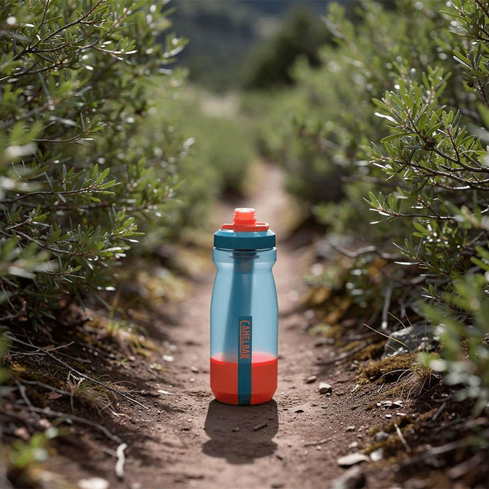 CamelBak Podium Bike Bottle 0.6L - Retail Colours - A drink bottle in turquoise and orange positioned on a dirt path surrounded by greenery, featuring a logo.