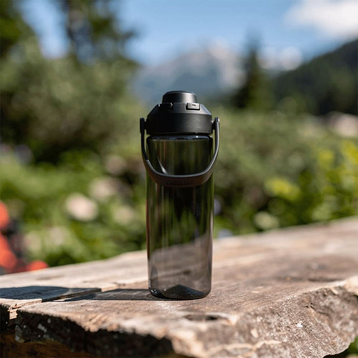 CamelBak - Thrive Chug 0.6L - A drink bottle in a sleek, transparent design with a black lid and handle, set against a natural outdoor backdrop.
