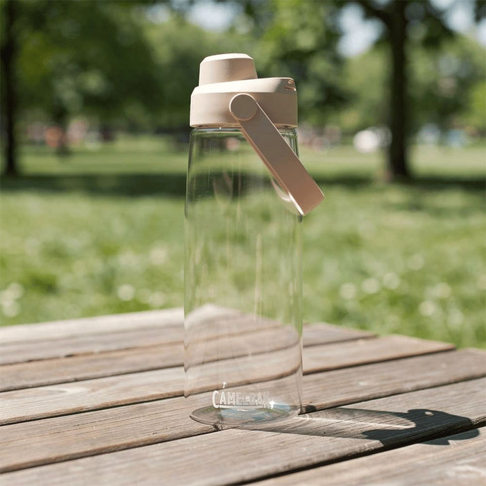 CamelBak - Thrive Chug 0.75L - A drink bottle with a clear body and a beige lid, featuring a handle, set on a wooden table in a grassy area.