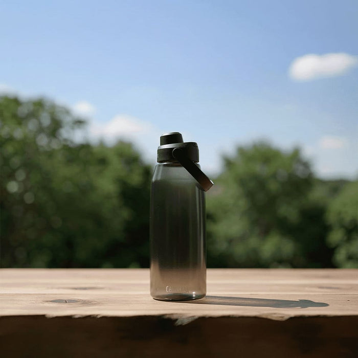 CamelBak - Thrive Chug 1.5L - A drink bottle in a gradient of grey with a black lid, placed on a wooden surface against a blurred green background.