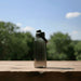 CamelBak - Thrive Chug 1.5L - A drink bottle in a gradient of grey with a black lid, placed on a wooden surface against a blurred green background.