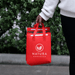 8L Insulated Reusable Bag - A red insulated reusable bag with two handles, featuring a logo on the front, held by a person.