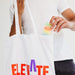Colourful Calico Tote Bag - A white tote bag with colourful lettering, being held by a person as they retrieve a clear container with coloured items.