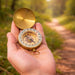 Luminous Compass - A brass compass in an open case, held in a hand, surrounded by a sunlit forest pathway.