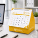 Desk Calendar With Organizer - A yellow desk calendar with an organiser, featuring a spiral binding and displayed on a workspace with a notebook and laptop.