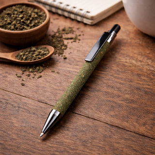 Tea Grounds Pen - A green pen with a silver tip rests on a wooden surface, surrounded by tea grounds and a notebook.
