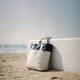 Field & Co.® Parks Day Tote - A beach tote bag in cream and navy rests against a white wall on sandy shore, with the ocean in the background.